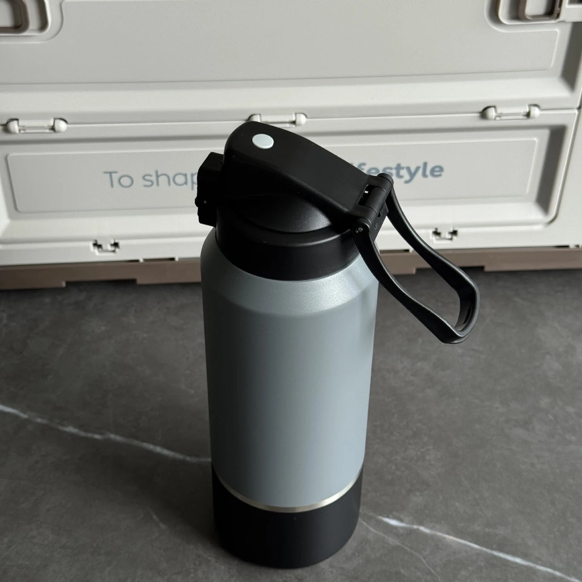 Gray insulated bottle with black lid and handle on a gray surface with a white container in the background.