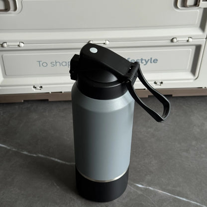 Gray insulated bottle with black lid and handle on a gray surface with a white container in the background.